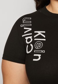 Černé tričko z bavlny s bílým vyšívaným textem umístěným vertikálně na rameni, který zní "Calvin Klein." Krátké rukávy.
