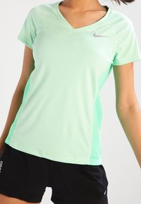 Mujer vistiendo una camiseta deportiva de manga corta verde claro y pantalones cortos negros, posando contra un fondo claro liso.