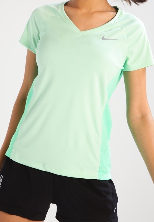 Mujer vistiendo una camiseta deportiva de manga corta verde claro y pantalones cortos negros, posando contra un fondo claro liso.