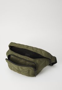 Bolso acolchado de color verde oliva con dos compartimentos con cremallera, textura suave, correa ajustable y diseño de patrón sutil.