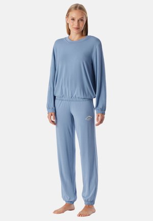 Frau steht barfuß und trägt ein locker sitzendes hellblaues Langarmshirt und passende Hosen mit elastischem Bund und einem kleinen Textlogo am linken Oberschenkel.