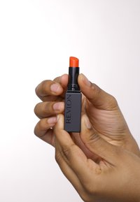 Rossetto Revlon in un elegante tubetto quadrato nero. Bullet arancione brillante con punta arrotondata. Mano che stringe il tubetto, evidenziando il suo design.