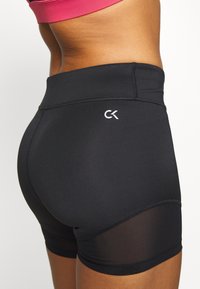 Musta, korkeavyötäröiset urheilushortsit, joissa on sileä pinta, läpinäkyvä paneeli ja valkoinen CK-logo sivussa.