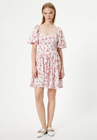 Bloemige korte jurk in lichtroze met pofmouwen, vierkante halslijn en geplooide rok. Kleine rode en groene bloemen creëren een zacht patroon.
