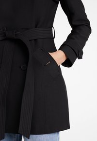 Ženský černý trench coat s páskem a knoflíkovými detaily, ruka v boční kapse, nošený přes modré džíny na bílém pozadí.