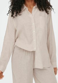 Camisa de lino color beige claro con botones, bolsillo en el pecho, mangas largas y corte relajado, combinada con pantalones de lino a juego.