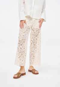 Pantaloni in pizzo color crema con un motivo a vortice floreale, design a gamba larga e orlo a scallop. Abbinati a sandali slide marroni.