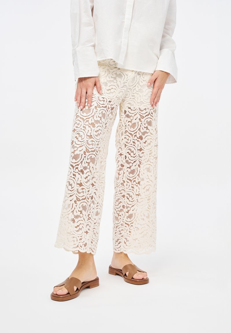 Pantaloni in pizzo color crema con un motivo a vortice floreale, design a gamba larga e orlo a scallop. Abbinati a sandali slide marroni.