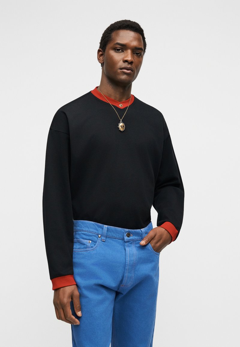 Jeune homme portant un sweat-shirt noir avec des poignets et un col rouges, un jean bleu et un collier avec pendentif, debout devant un fond gris uni.