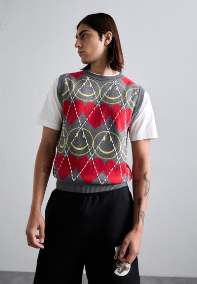 Junge Person mit schulterlangem Haar trägt einen grauen ärmellosen Pullover mit roten Herzen und gelben Smiley-Gesichtern über einem weißen T-Shirt.
