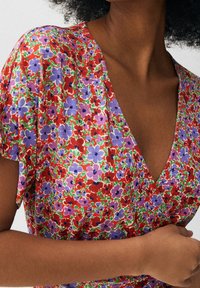 Robe fleurie avec motifs rouges, violets et verts, en tissu léger, col en V et manches courtes. Gros plan sur les détails du col et de l'épaule.