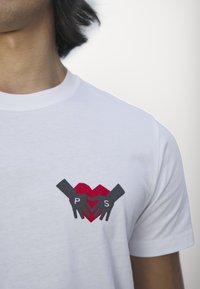 White cotton t-shirt featuring a graphic of two hands holding a heart, with red and grey colors, and the letters "P S" in black on the heart.