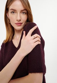 Bordeaux gebreide trui met een v-hals, korte mouwen en een zachte textuur. Model draagt een delicate ring op haar hand met ingewikkelde ontwerpdetails.