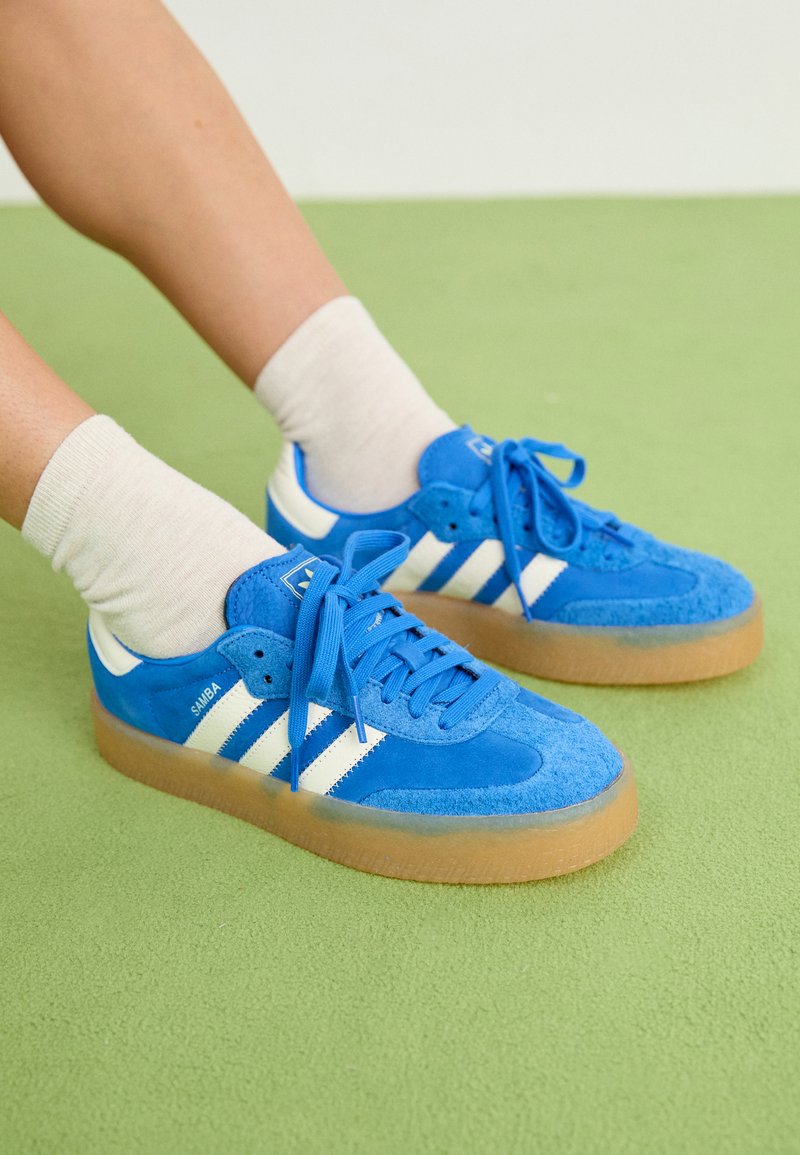 Blå semskede sneakers med hvite striper, designet med en gummisåle, lisser og en polstret krage. Myk tekstur, atletisk stil.