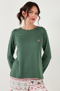 Mujer con cabello castaño recogido en una coleta, vistiendo una camiseta de manga larga verde bosque con un pequeño bordado floral y pantalones con un patrón festivo.