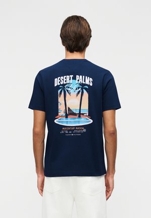 Mann trägt ein marineblaues T-Shirt mit einem Midcentury-Haus, Palmen und Pool-Grafik auf dem Rücken, kombiniert mit weißen Hosen.