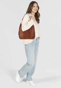Borsa a spalla in pelle intrecciata marrone con texture a rete, forma arrotondata e una sola tracolla. Abbinata a un maglione crema e jeans azzurri chiari.