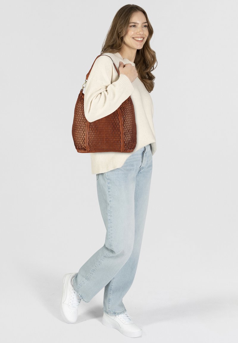 Borsa a spalla in pelle intrecciata marrone con texture a rete, forma arrotondata e una sola tracolla. Abbinata a un maglione crema e jeans azzurri chiari.
