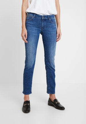 Kvinde iført blå slim-fit jeans, hvid top og sorte læder loafers med guldspænder, stående på en hvid gulv og baggrund.