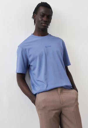 Mężczyzna opierający się o białą ścianę, ubrany w jasnoniebieską koszulkę BOSS oraz jasnobrązowe spodnie, ręce w kieszeniach, zrelaksowana postura.