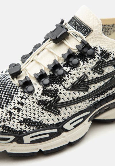 Κοντινό πλάνο μαύρου και άσπρου πλεκτού αθλητικού παπουτσιού με ελαστικά κορδόνια χωρίς δέσιμο, στερεωμένα με μαύρα πλαστικά κλιπ και διακοσμητικά πλευρικά στοιχεία σε σχήμα βέλους.