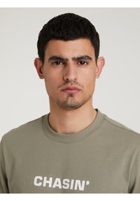 Olivazöld pulóver bordás nyakkal, az elején merész fehér "CHASIN'" felirattal. Simább textúra, laza fazon.