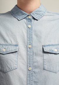 Lysblå denimskjorte med knaplukning foran, spids krave, to brystlommer og synlige syninger.