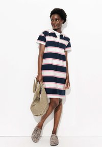 Femme portant une robe polo rayée bleu marine, blanche et rouge, tenant un sac beige, avec des chaussures à enfiler à imprimé léopard, se tenant contre un mur blanc.