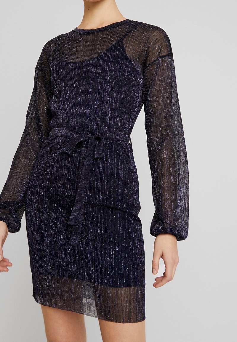 Robe violette scintillante en matériau transparent et texturé. Manches longues et silhouette ajustée, avec une ceinture à nouer à la taille.
