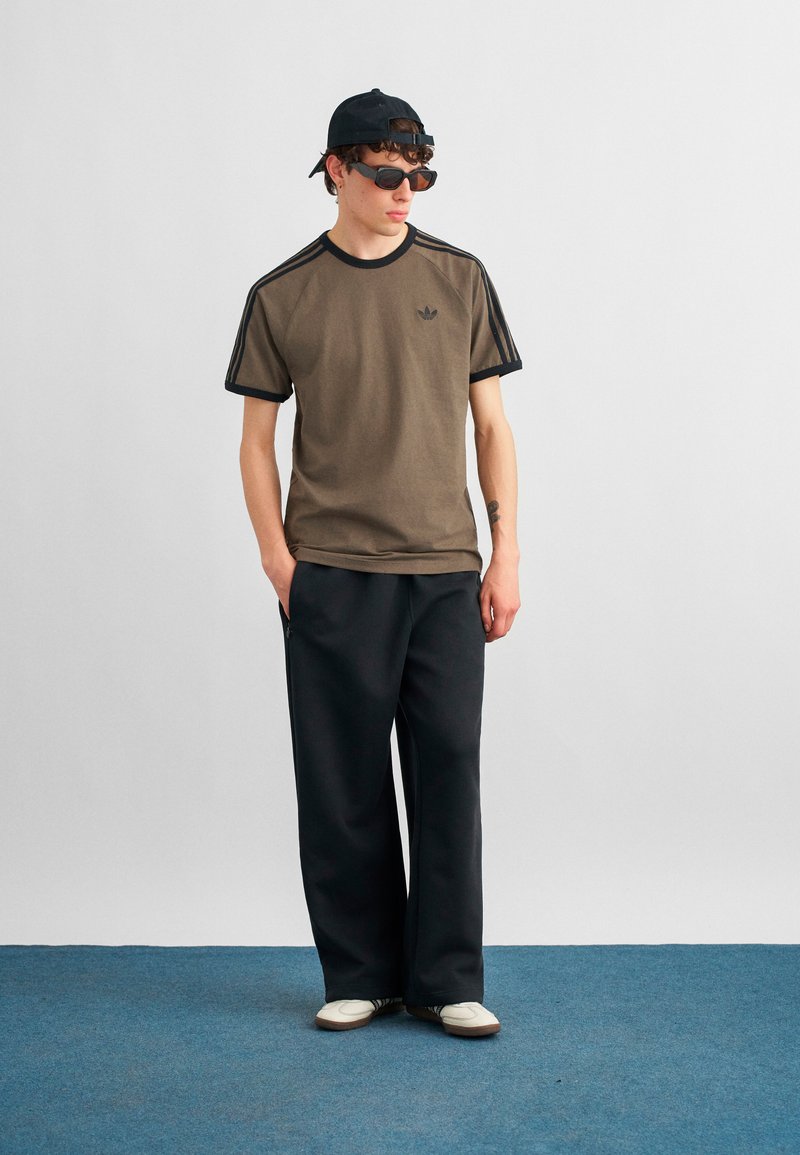 Ung mand iført brun Adidas t-shirt, sorte bukser med vide ben, hvide sneakers, sort kasket bagudvendt og mørke solbriller, stående på blåt tæppe.