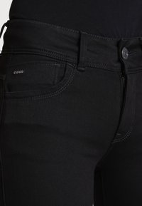 Jeans de mezclilla negra con un ajuste slim, que cuentan con un botón de metal, cremallera y costuras visibles. Incluye un bolsillo frontal con un detalle de logo.