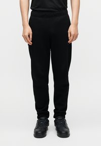 Μαύροι φόρμες με ελαστική ζώνη μέσης, στενά πέδια και πλαϊνές τσέπες, συνδυασμένοι με μαύρα αθλητικά παπούτσια, στέκεται σε απλό φόντο.