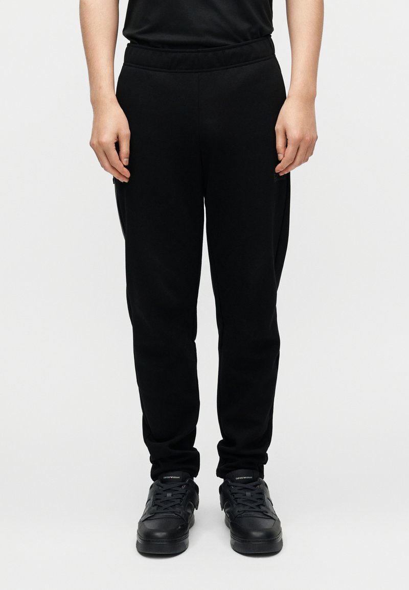 Μαύροι φόρμες με ελαστική ζώνη μέσης, στενά πέδια και πλαϊνές τσέπες, συνδυασμένοι με μαύρα αθλητικά παπούτσια, στέκεται σε απλό φόντο.