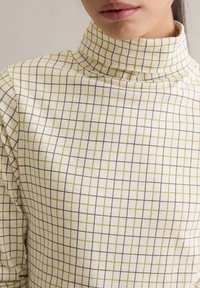 Crèmekleurige coltrui met een ruitpatroon in donkergrijs en olijfgroen, gemaakt van zacht katoenmateriaal. Close-up van de stoftextuur.