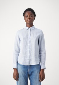 Jasnoniebieska koszula z długim rękawem zapinana na guziki, wykonana z teksturowanego lnu, z klasycznym kołnierzykiem i lekko zaokrąglonym dołem. Nosi ją z niebieskimi dżinsami.