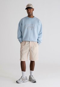 Sudadera azul claro con el logo "Prohibido", pantalones cortos beige con pliegues y zapatillas deportivas grises. Gorra con rayas y visera en contraste.