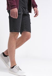 Svarta denimshorts med en distressed design och fransade kanter, tillsammans med ljusgrå sneakers och en vinröd zip-up jacka.