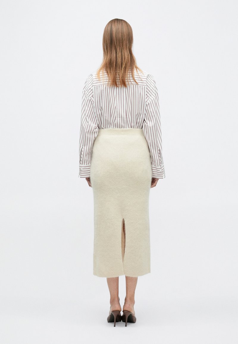 Woman standing facing away, wearing a white striped shirt, cream fuzzy midi skirt with back slit, and dark high-heeled shoes.