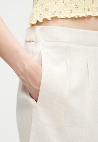 Main reposant dans la poche d'un pantalon taille haute beige clair texturé, associé à un haut court en tricot jaune.