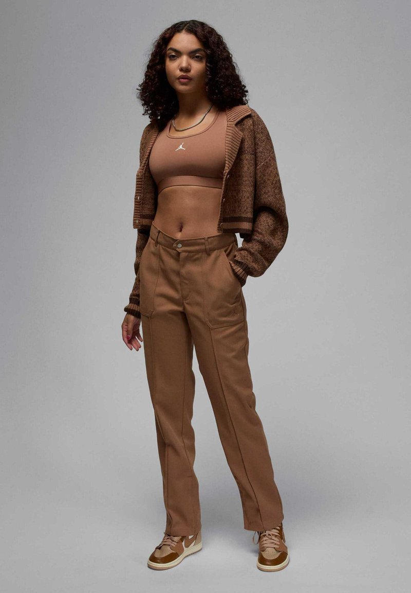 Cardigan corto lavorato a maglia marrone sopra un reggiseno sportivo beige, abbinato a pantaloni alti marroni e sneakers color cammello. Design minimalista.