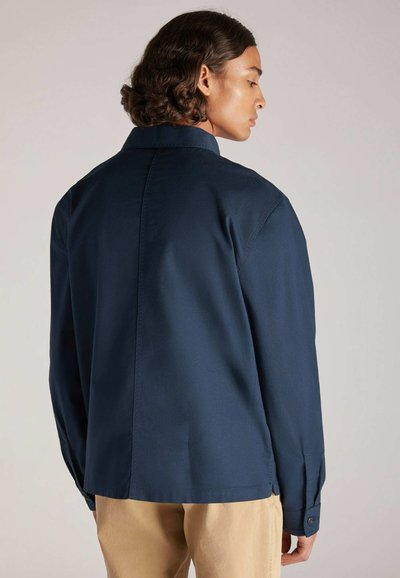 Un giovane in una camicia blu navy si volta di spalle, mostrando il retro della camicia e i suoi capelli ricci. Lo sfondo è semplice e neutro.