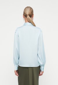 Blusa de manga longa azul clara com colarinho, detalhes franzidos nos ombros, textura suave e punhos com botões, combinada com uma saia verde oliva.