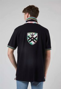 Polo noir avec un écusson coloré brodé au dos, un col à motif et des accents de manches rayés, fabriqué en tissu doux.