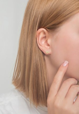 Goldfarbene, schlangenförmige Ohrstecker mit glänzender Oberfläche, die an einem Ohr eines Models getragen werden, während ein neutraler Finger auf das Ohr zeigt.