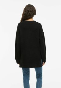 Pulôver preto de malha oversized com mangas longas, decote redondo e um tecido texturizado. Usado com jeans azuis de perna reta.