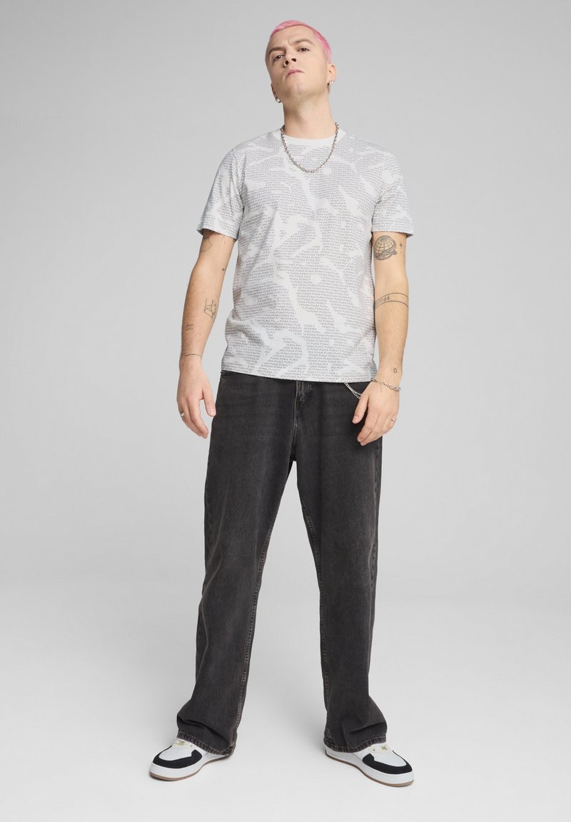 Grå camouflage-mønstret t-shirt med korte ærmer, parret med mørkeblå, løstsittende jeans og sort-hvide sneakers. Tyk sølvkæde om halsen.