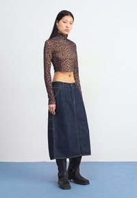 Model står på blå gulv iført en tætsiddende leopardprint top, et denimskørt og sorte ankelstøvler, mod en helt hvid baggrund.