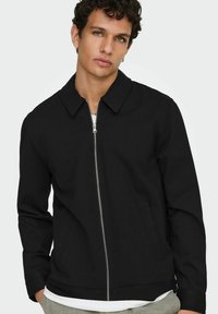 Ung mann med krøllete hår iført en svart jakke med glidelås over en hvit skjorte og grå bukser, stående mot en ensfarget lys bakgrunn.