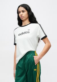 White cotton t-shirt with black ringer neckline, featuring "adidas" logo. Paired with green track pants with yellow stripes and logo accents.