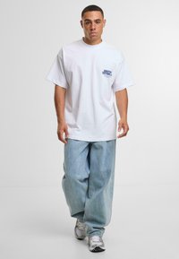 Valkoinen puuvillapaita, jossa on sinistä tekstiä vasemmassa rintataskussa. Yhdistetty vaaleansinisiin farkkuihin ja harmaisiin urheilukenkiin. Rennonistuva.